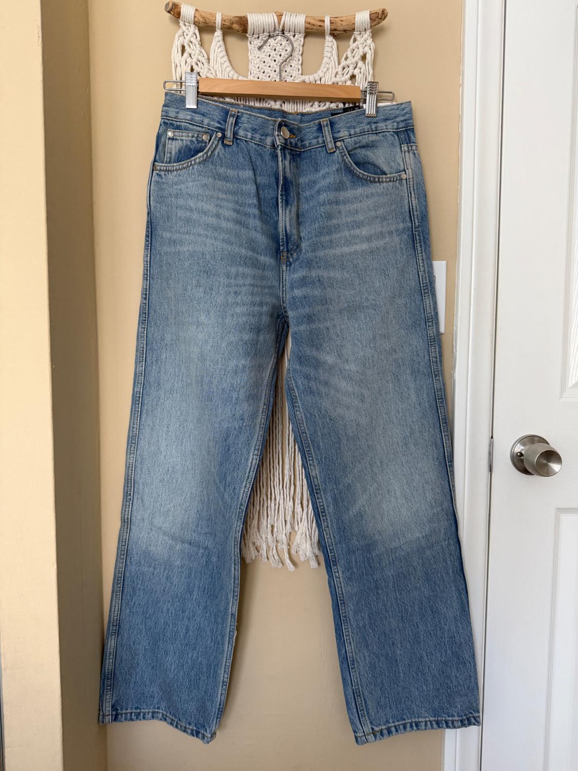 Wide-leg blue jeans hanging on a wooden hanger against a beige background with macrame details.