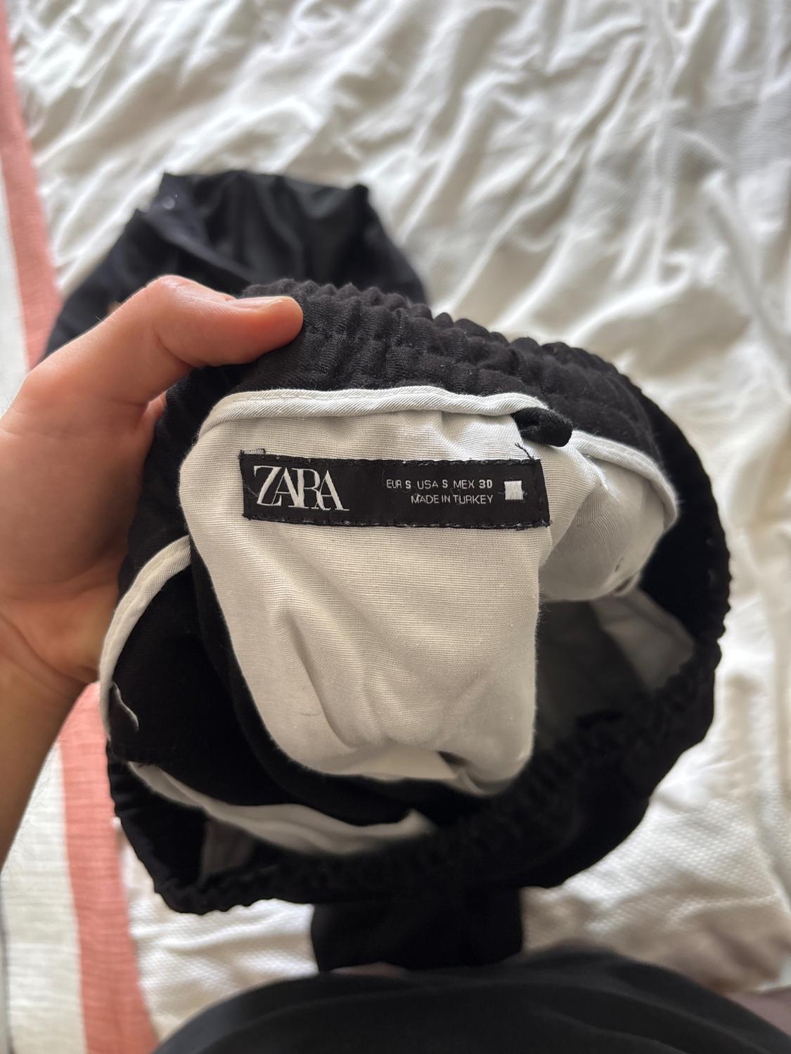 Hand holding a black garment with an elasticated waist against white sheets. Text: ZARA EUR S USA S MEX 30 MADE IN TURKEY.