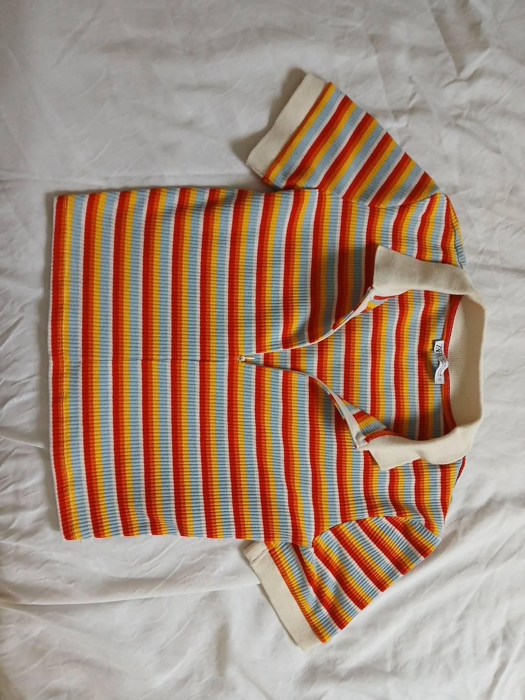 Ribbed polo shirt with orange, red, yellow and blue stripes featuring a cream collar, zip fastening and ZARA label, on crumpled white fabric.