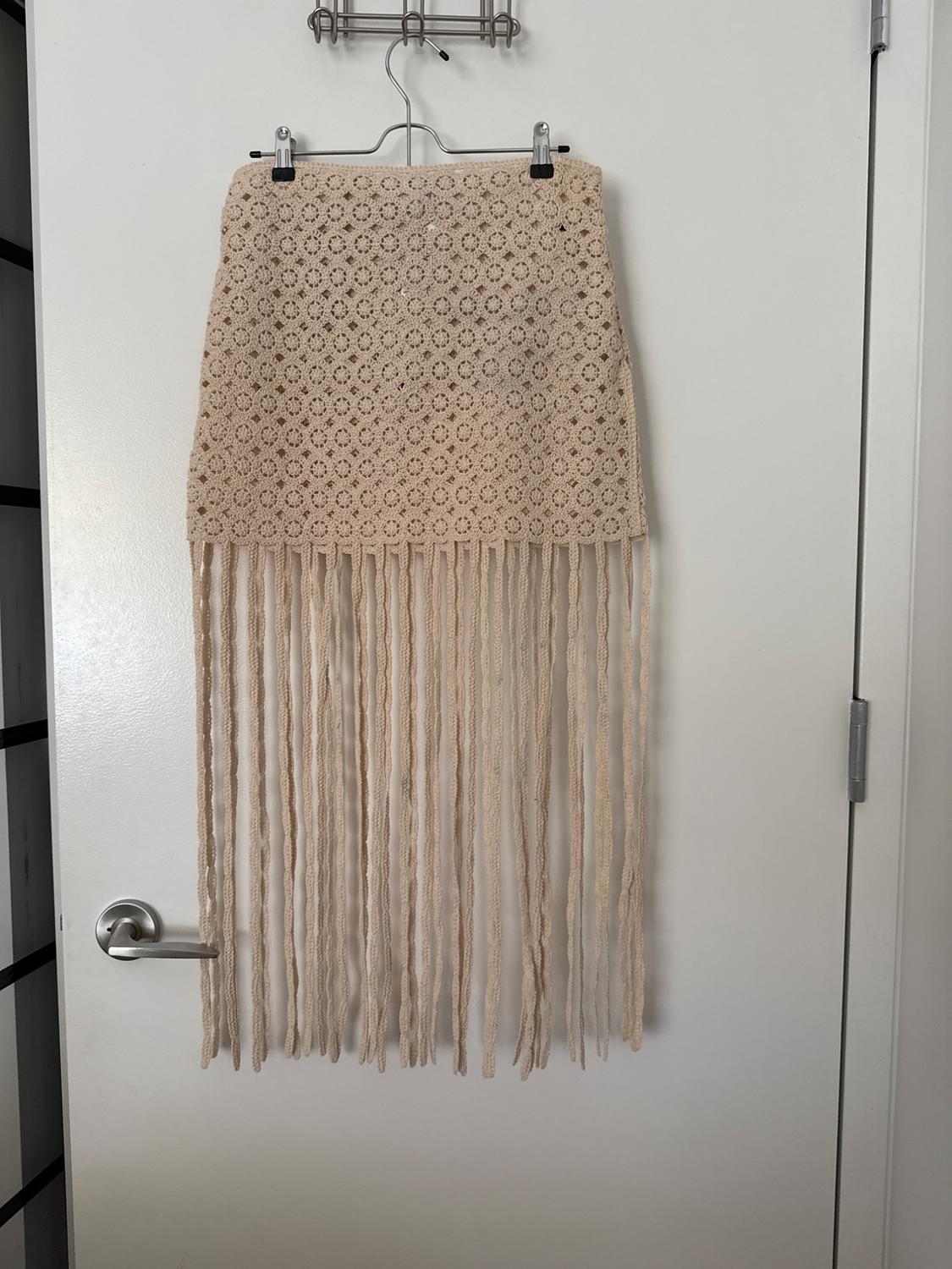 Beige crochet skirt with long fringing, hanging on a metal hanger on a white door. Bohemian style and relaxed vibe.