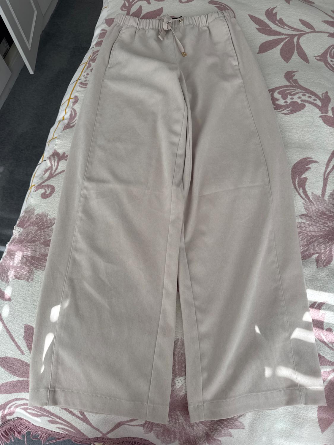 Wide-leg beige trousers with an elasticated drawstring waist, on a bed with a pink floral bedspread and a grey background.
