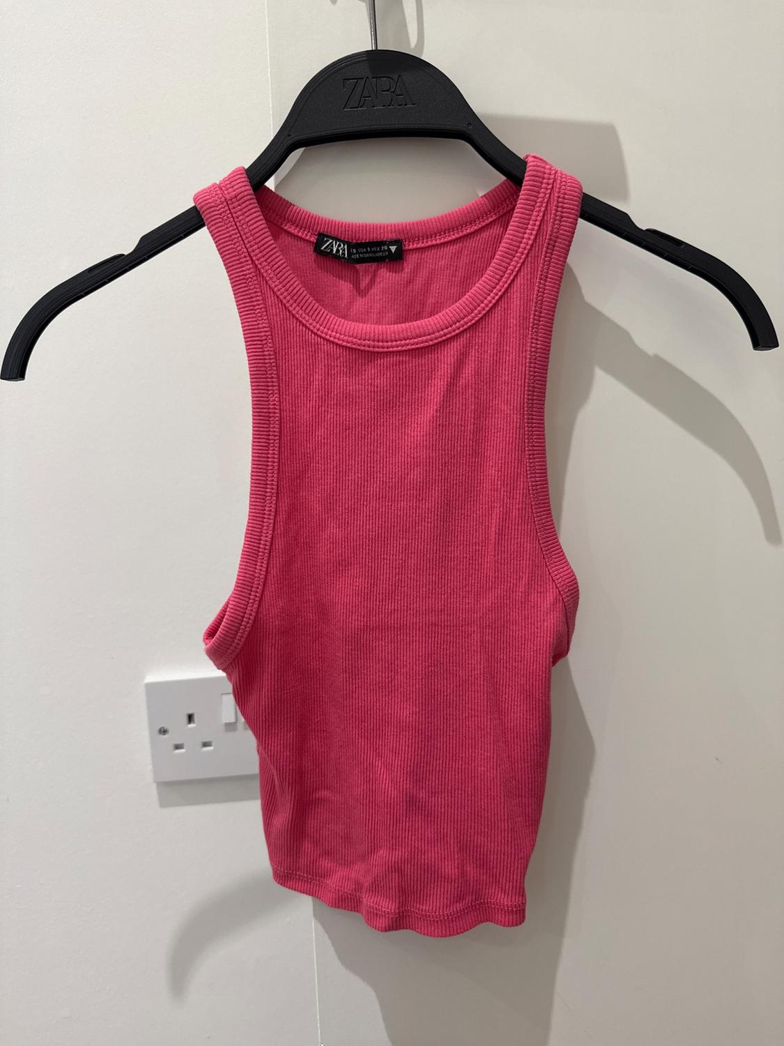 Pink sleeveless top hanging on a black hanger with the brand visible. White background with a plug socket on the wall.