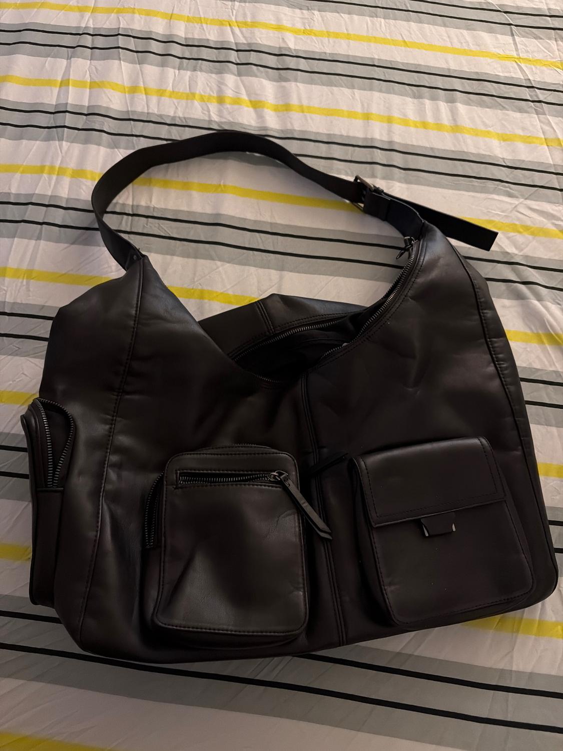 Large black bag with multiple pockets and straps, on a grey and yellow striped quilt.