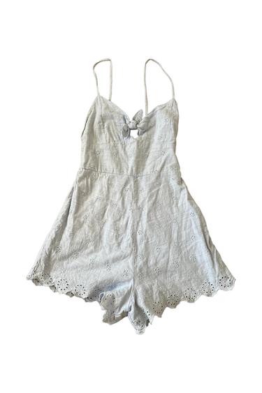 Light blue strappy romper featuring a front tie, keyhole cutout, eyelet details, and a scalloped hem on a white background.