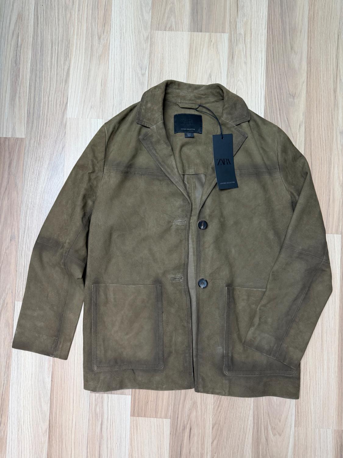 Brown suede jacket with two front pockets and black ZARA label, on a light wooden floor.