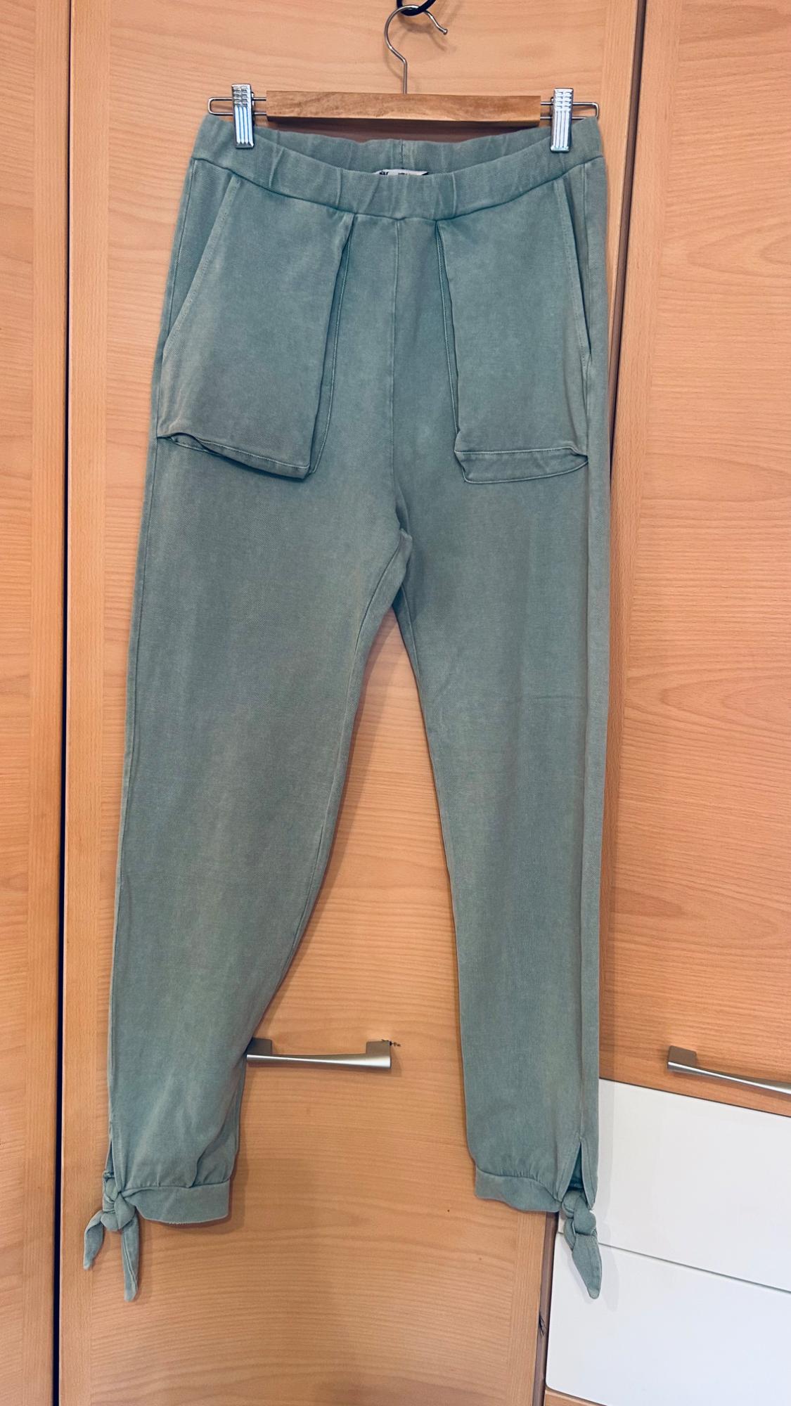 Light green casual trousers with an elasticated waist, large pockets and ties at the hems, hanging from a hanger in a wooden wardrobe.