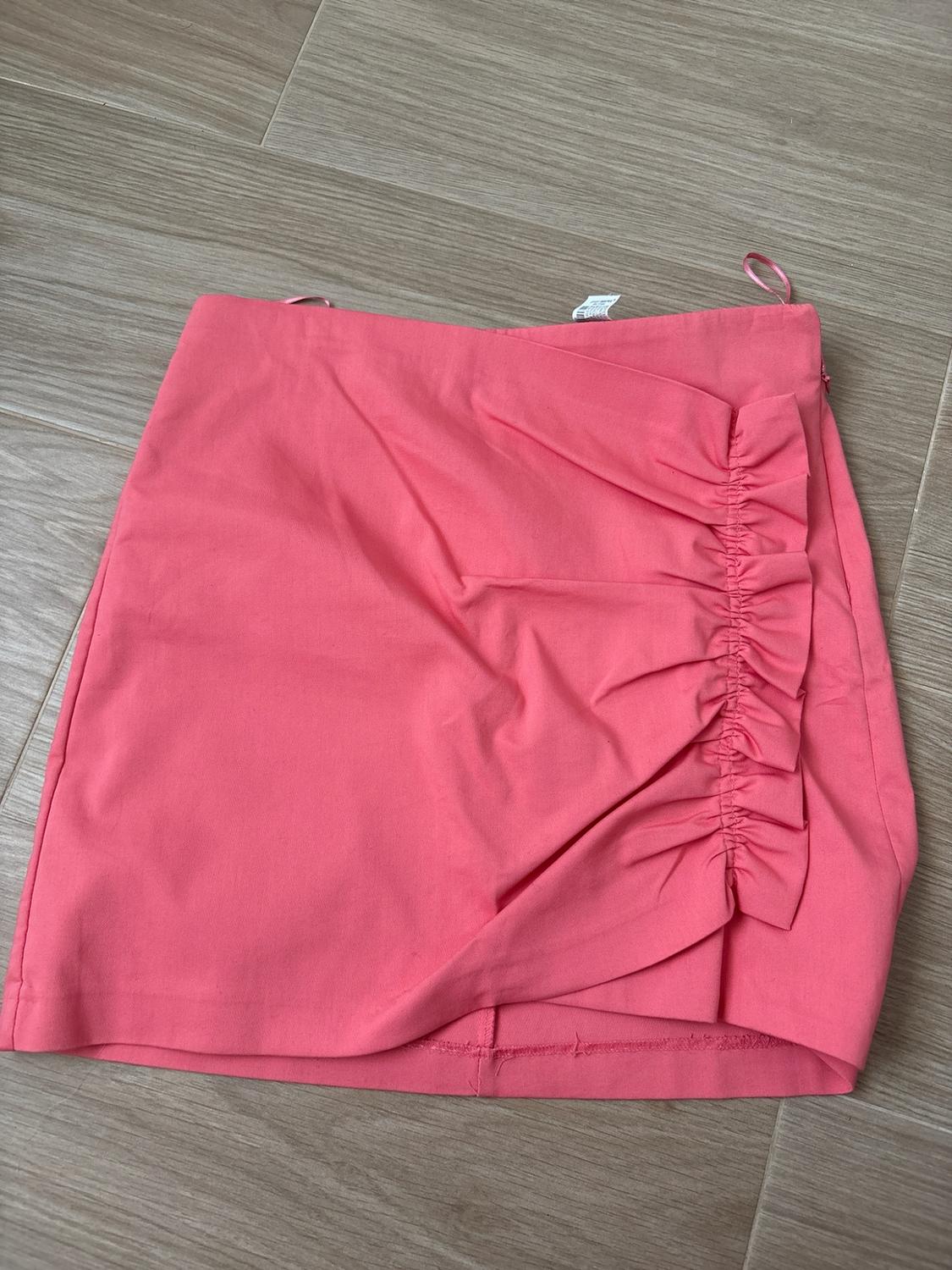 Pink mini skirt with side ruching detail, laid out on a light wood floor. Simple composition and casual style.