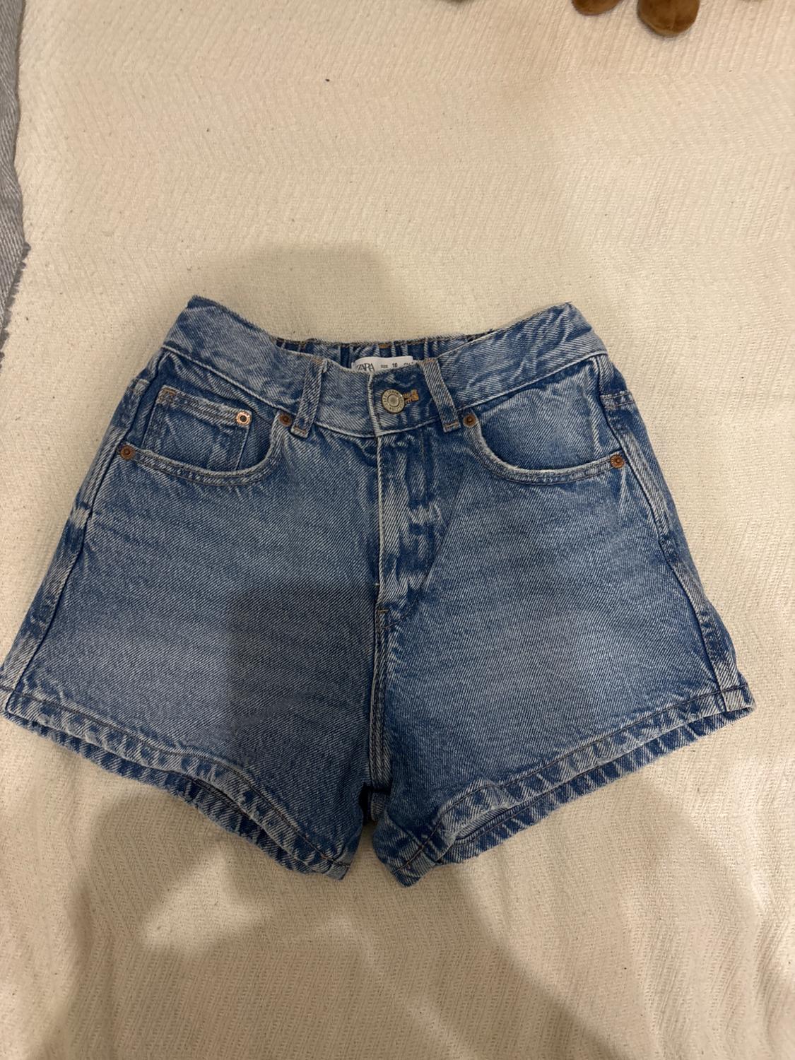 Light blue denim shorts with five pockets and button fastening, on a textured beige surface.