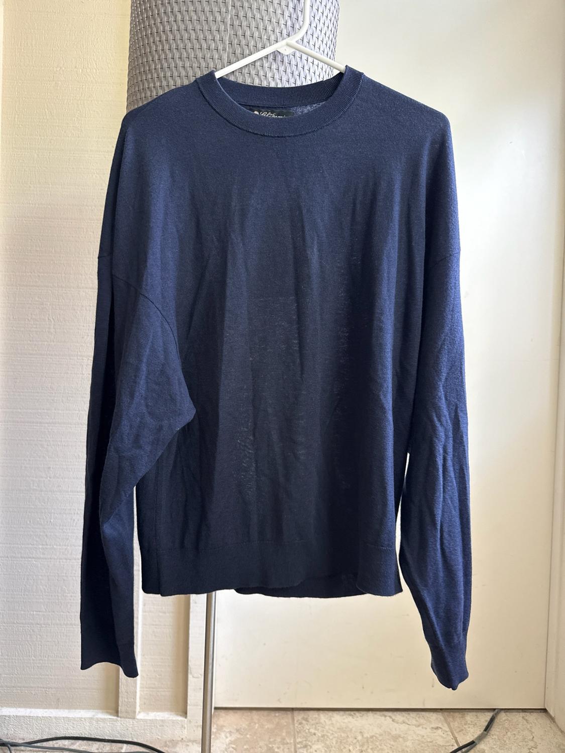 Dark blue long-sleeved jumper hanging on a white hanger on a metal stand, against a light-coloured wall.