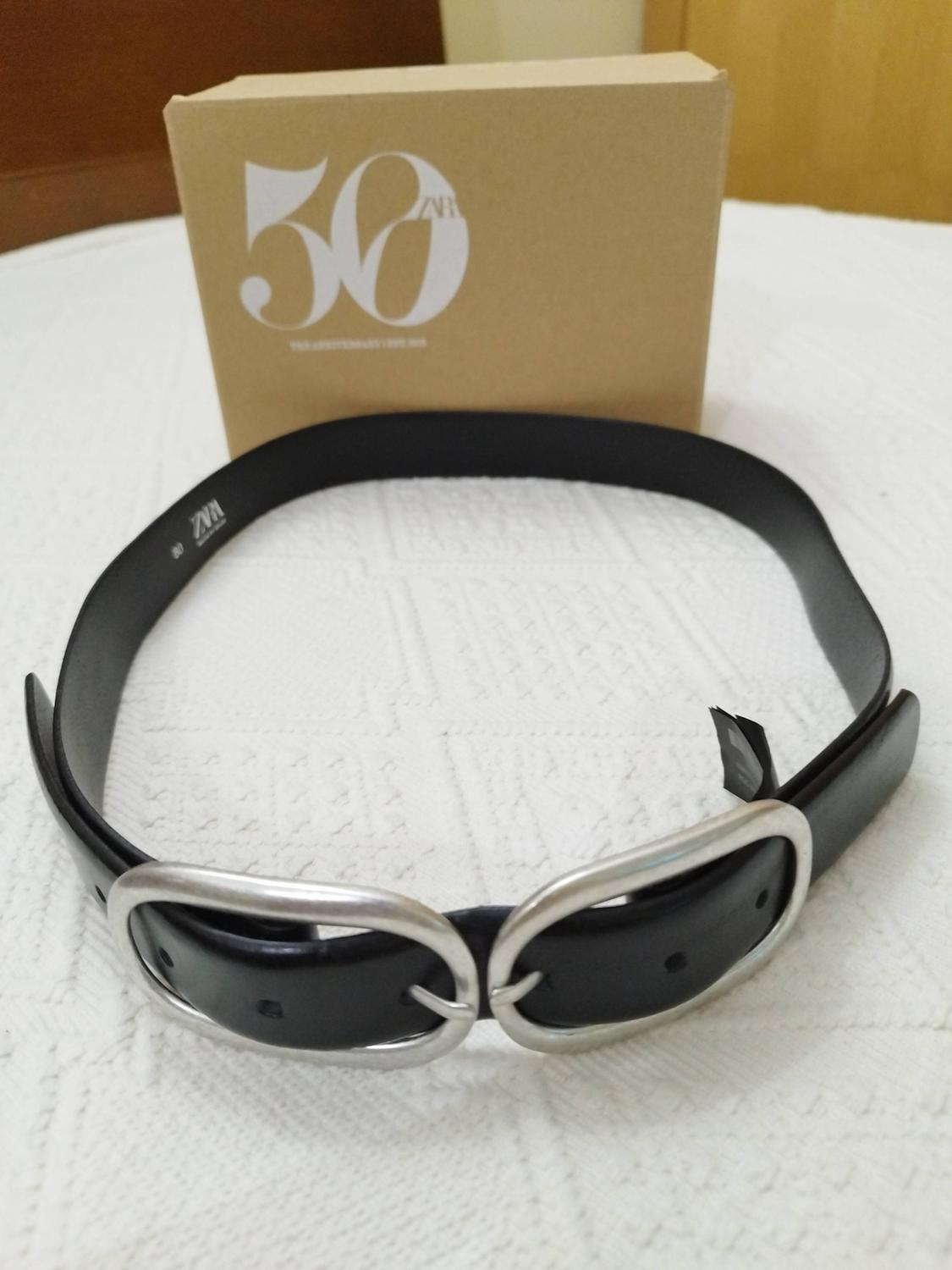 Black belt with two oval silver buckles on white fabric. In the background, a cardboard box with the text "50 ZARA".
