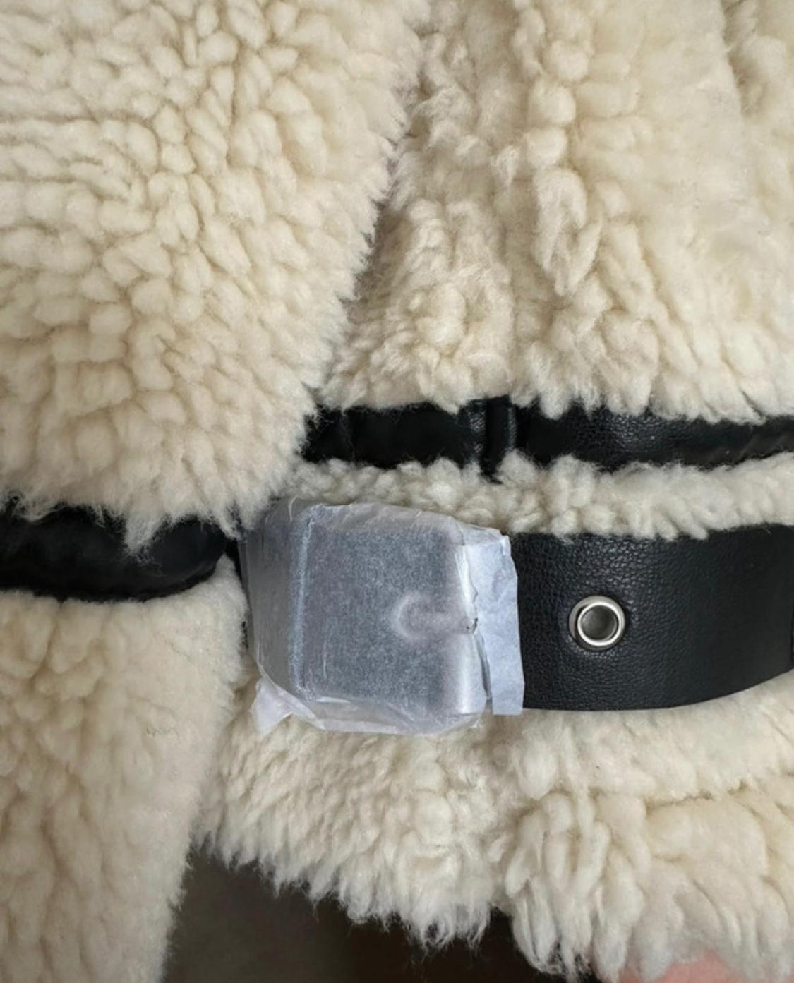 Detail of a raw curly-haired garment with black faux-leather straps and a metal eyelet. The buckle is covered with protective paper.