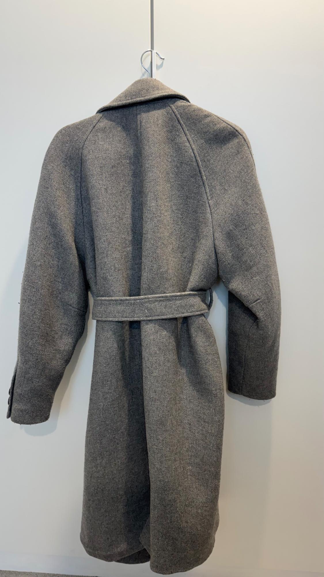 Grey wool coat hanging on a hanger, seen from the back, with a matching belt and a white background.