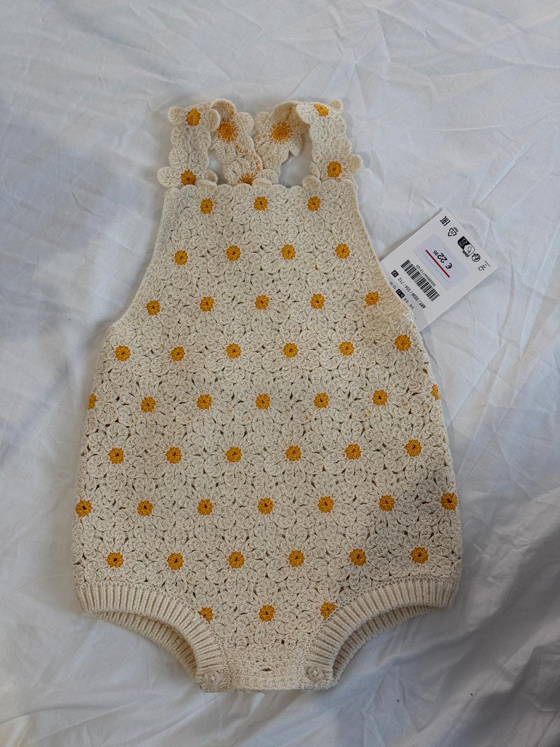 Beige crochet romper with yellow flowers on a white background. Visible tag with barcode and price of €22.