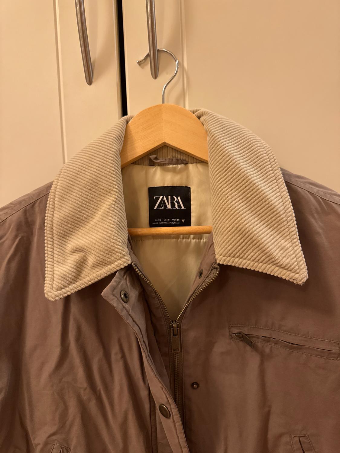 Brown jacket with beige corduroy collar hanging on a wooden hanger. Visible label with the text "ZARA". Light wardrobe background.