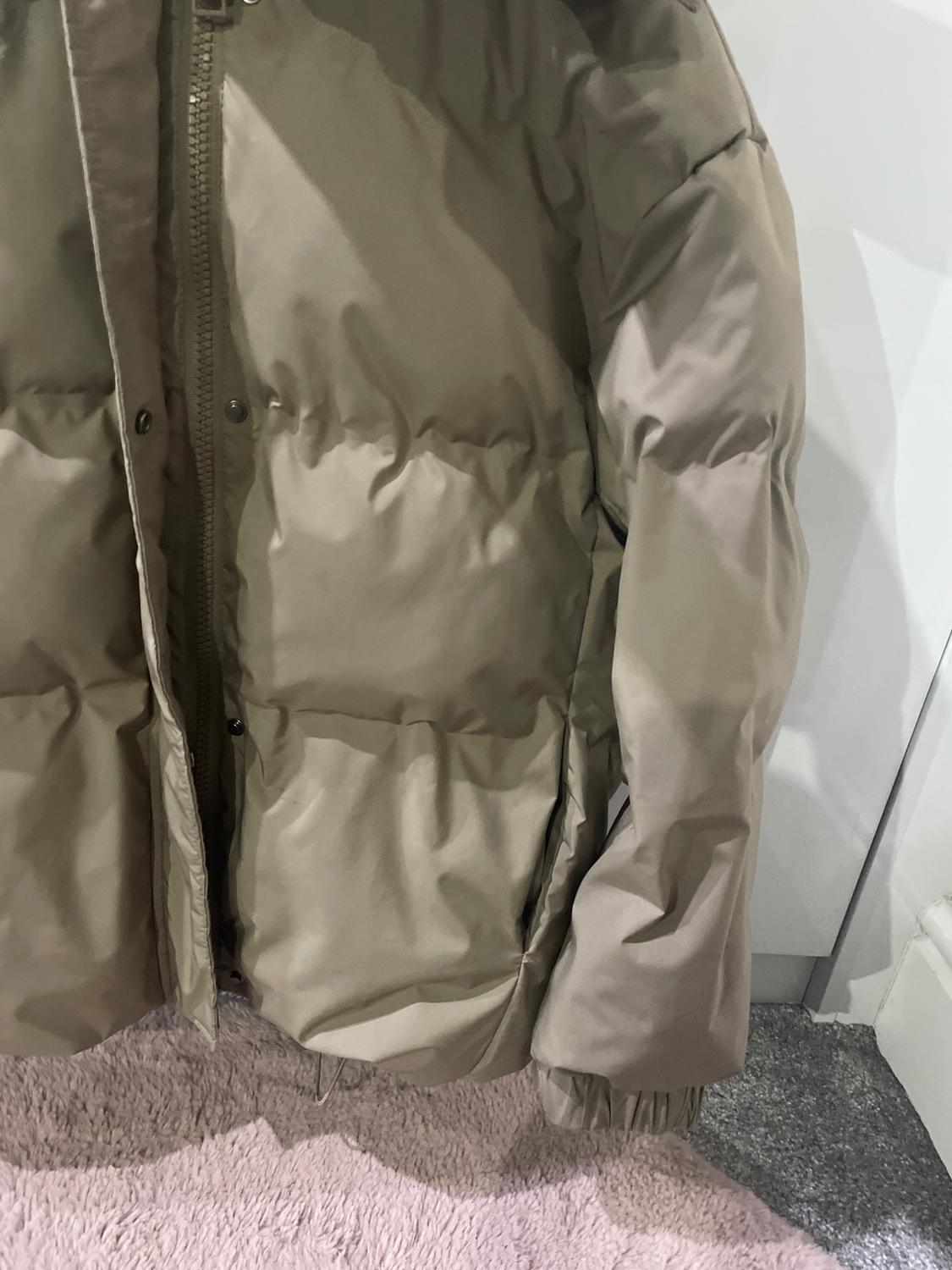 Beige quilted jacket hung over a pink rug, with visible zip and press studs. White and grey background.