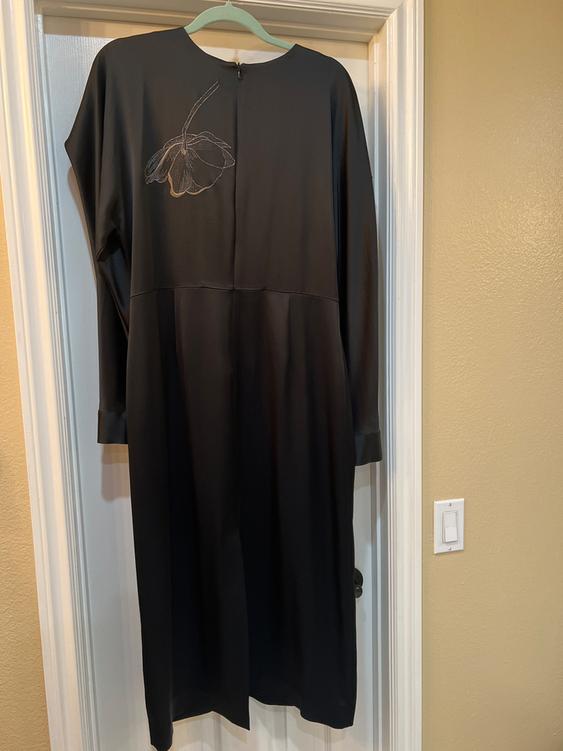 Black dress hanging on a door, with embroidered flower design on the left shoulder. Interior setting with a beige wall.