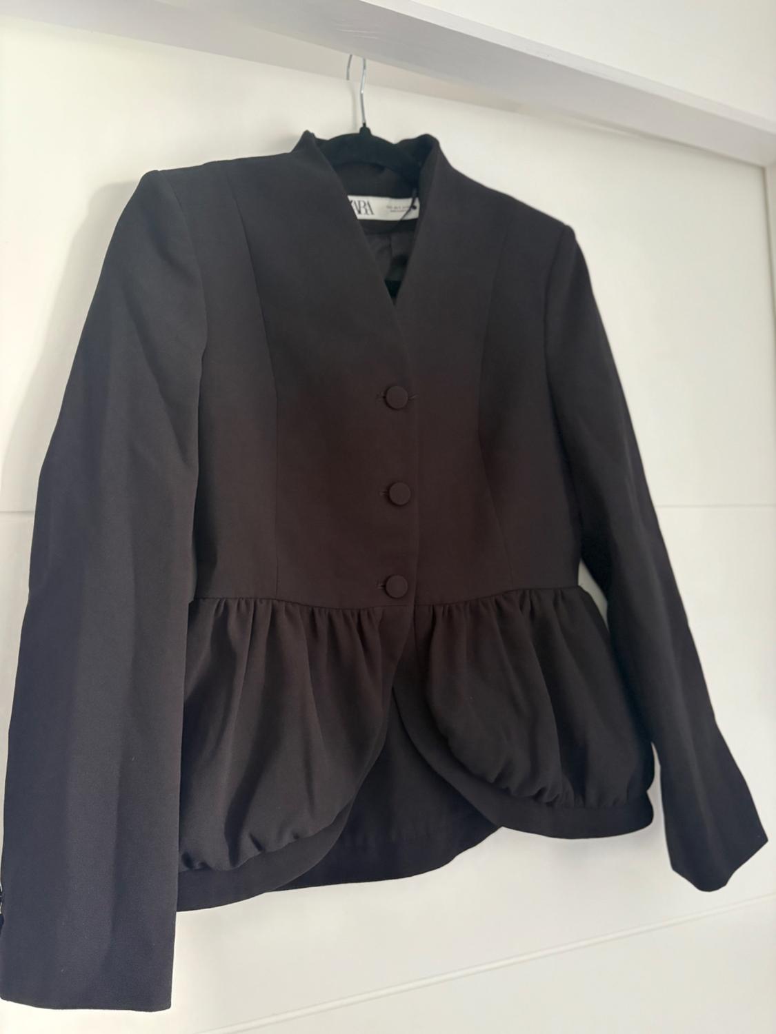Black jacket with buttons and a pleated waist design, hanging on a hanger against a white background.