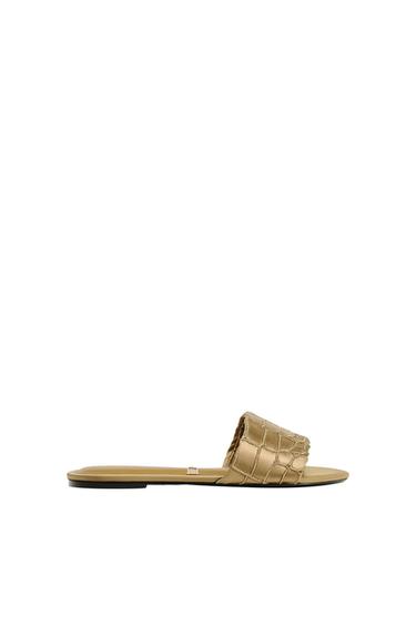Gold flat sandal with a textured design on the wide strap, on a white background.