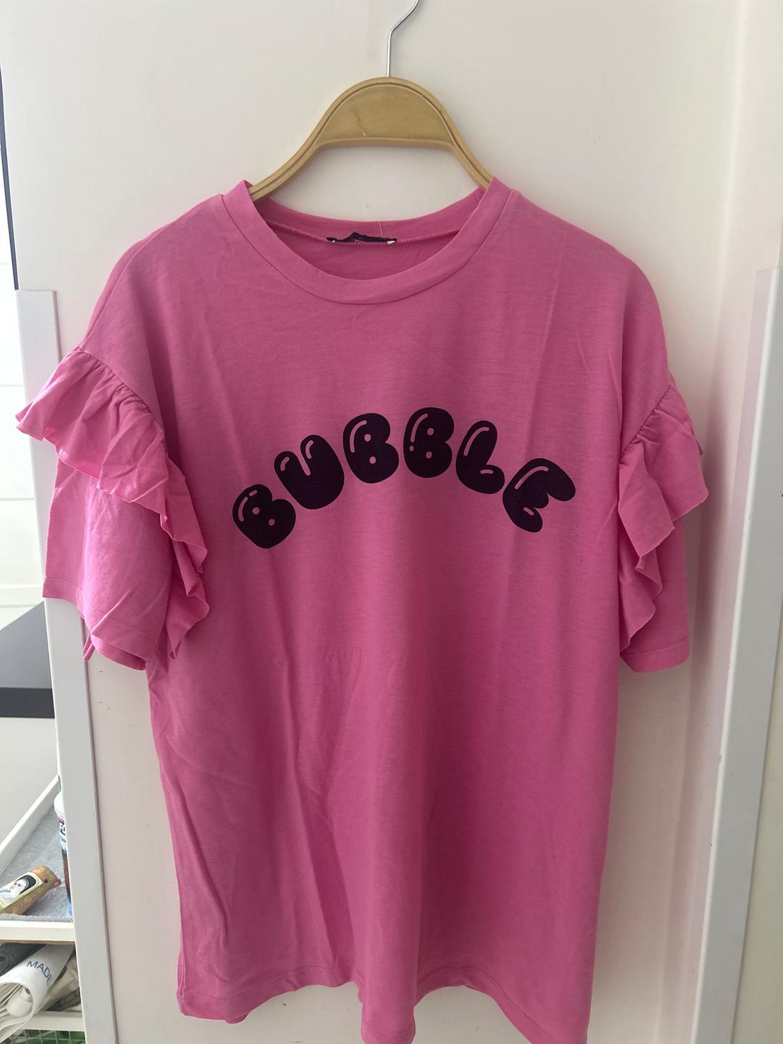 Pink T-shirt with ruffled sleeves and the word "BUBBLE" in black letters on the chest, hanging on a wooden hanger.