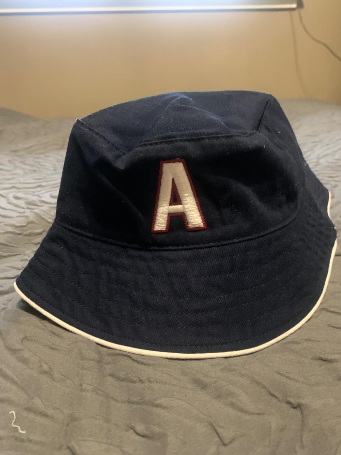 Dark blue hat with a white brim and the letter "A" embroidered on the front, on a grey bed. Soft light comes in through the window.