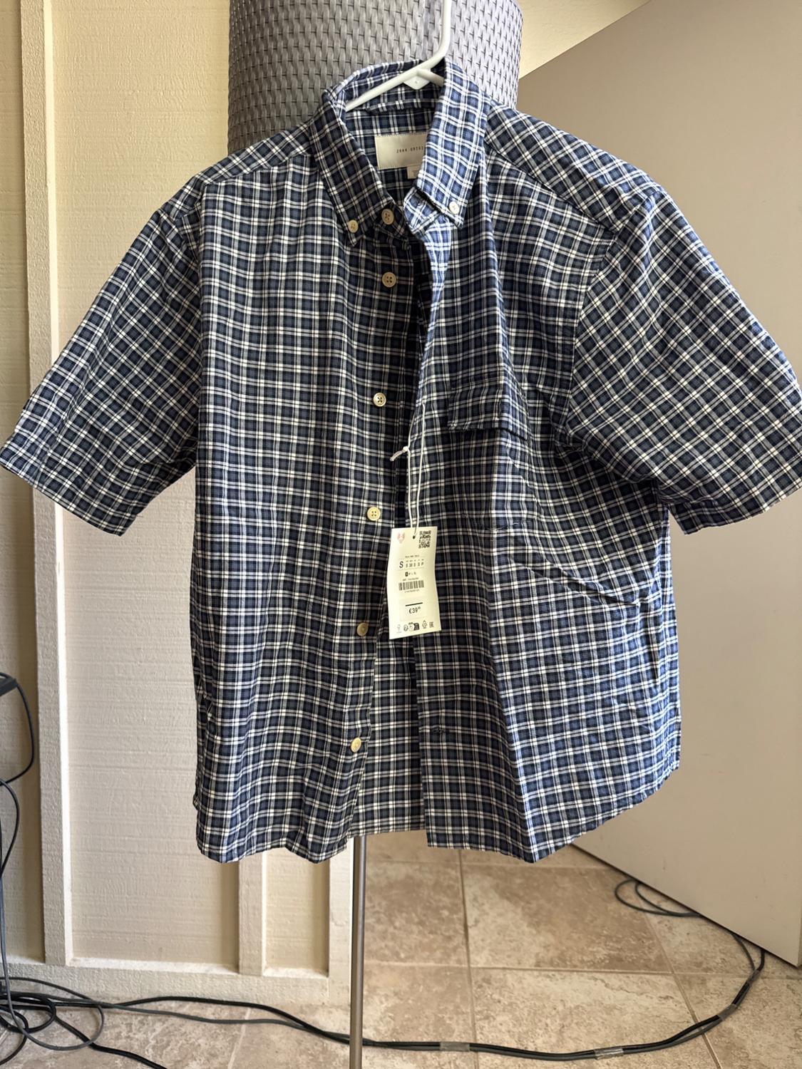 Short-sleeved blue and white checked shirt on a hanger. Text: ZARA ORIGINS S €39. Indoor setting with tiled floor and cables.