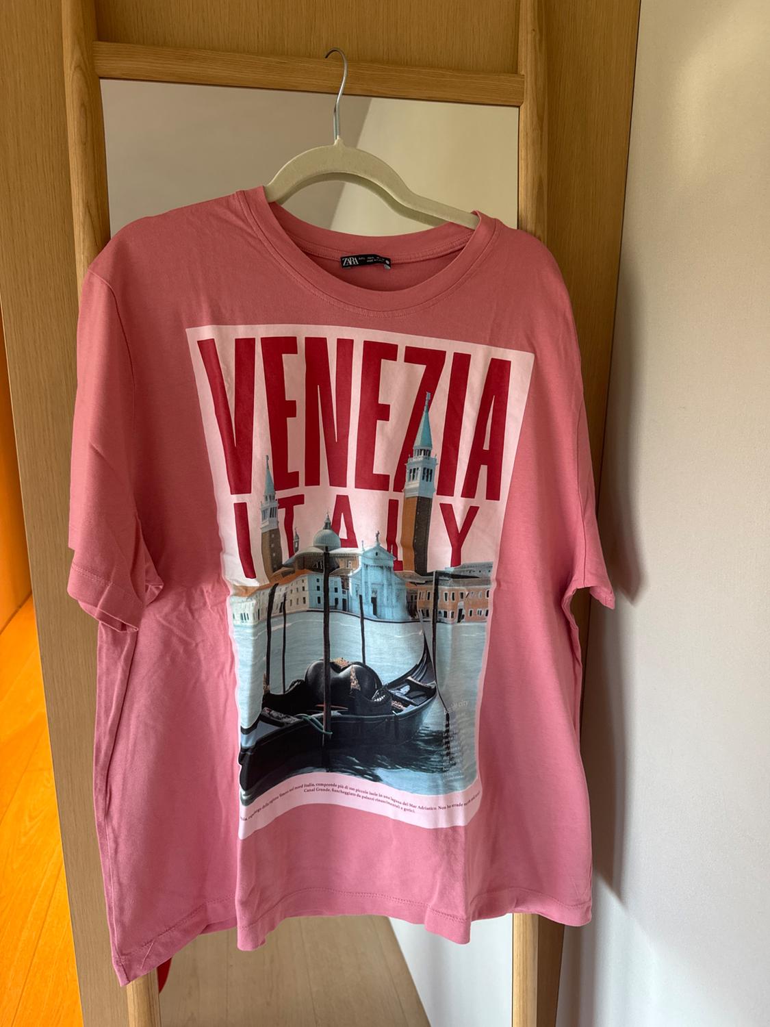 Pink t-shirt hanging on a mirror, with an image of Venice and the text "VENEZIA ITALY" in large red letters.
