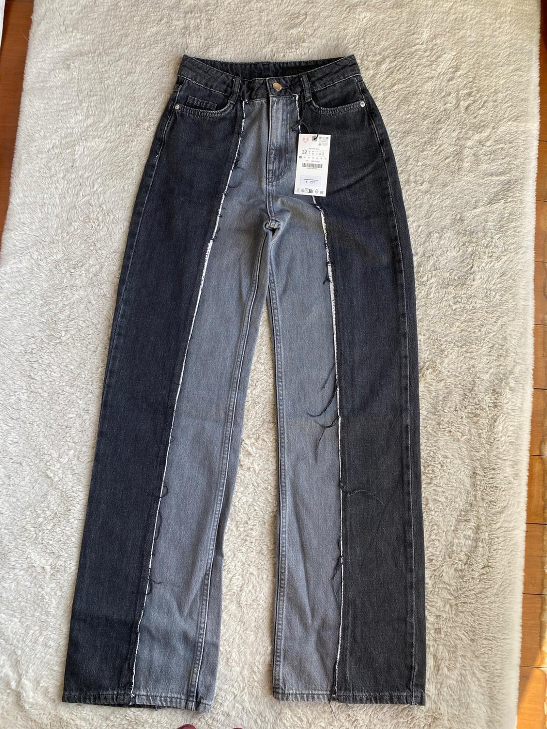 Straight-leg jeans in two shades of grey and black, with frayed stitching and a label bearing the text “32”, on a white rug.