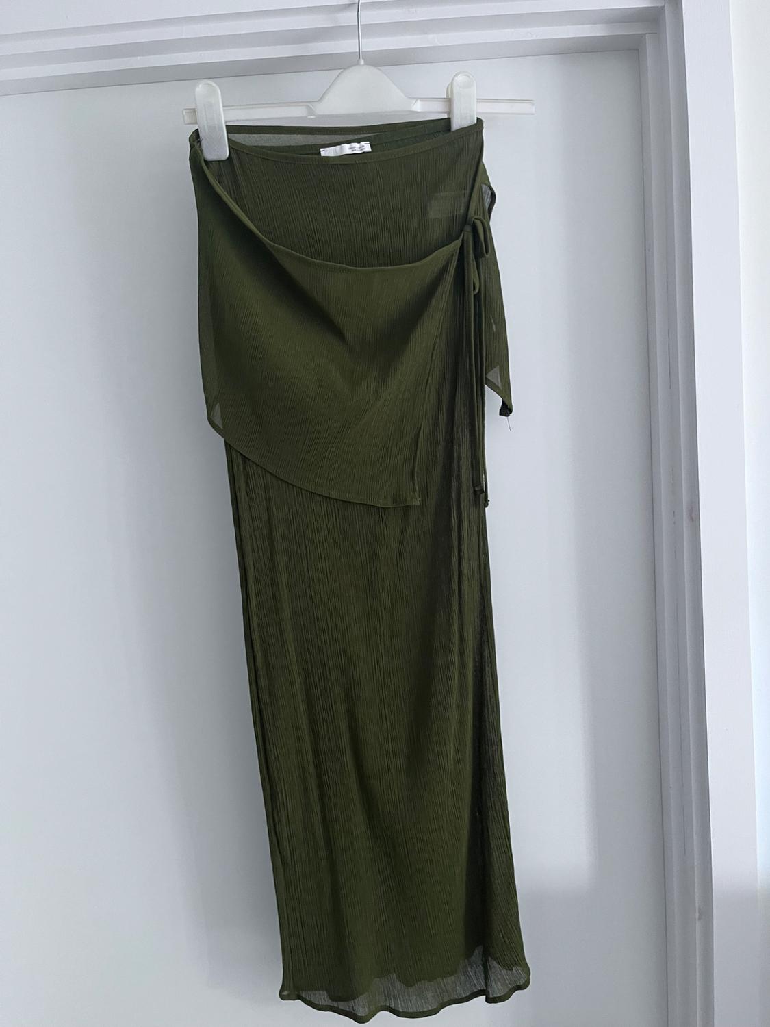 Long olive green skirt in textured fabric with draped detail and side tie, hanging on a white hanger in front of a door.