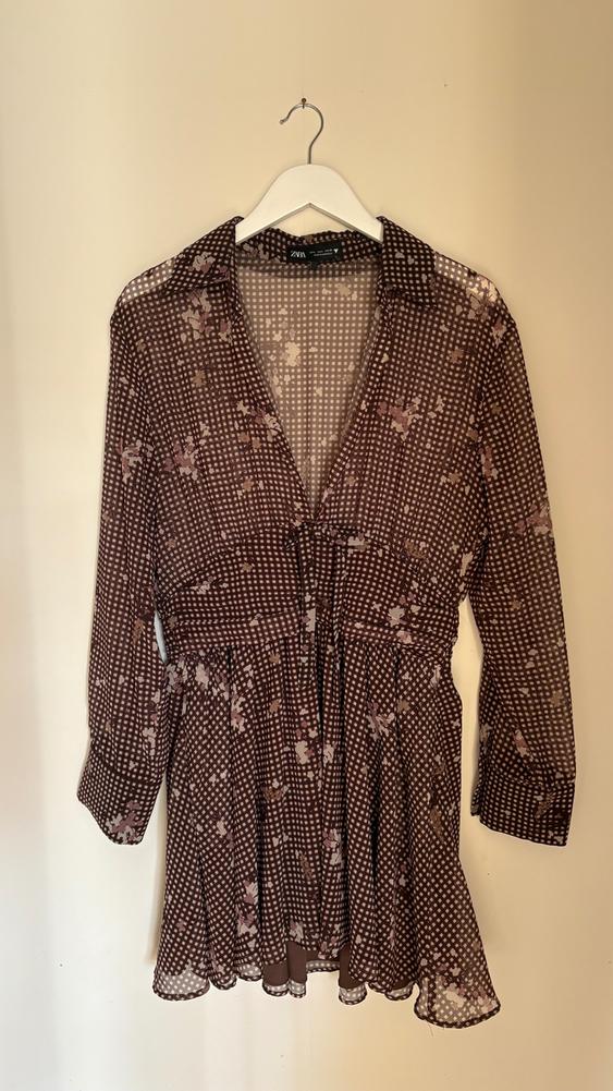 Brown dress with a checked and floral print, long sleeves, hanging on a white hanger against a beige background.