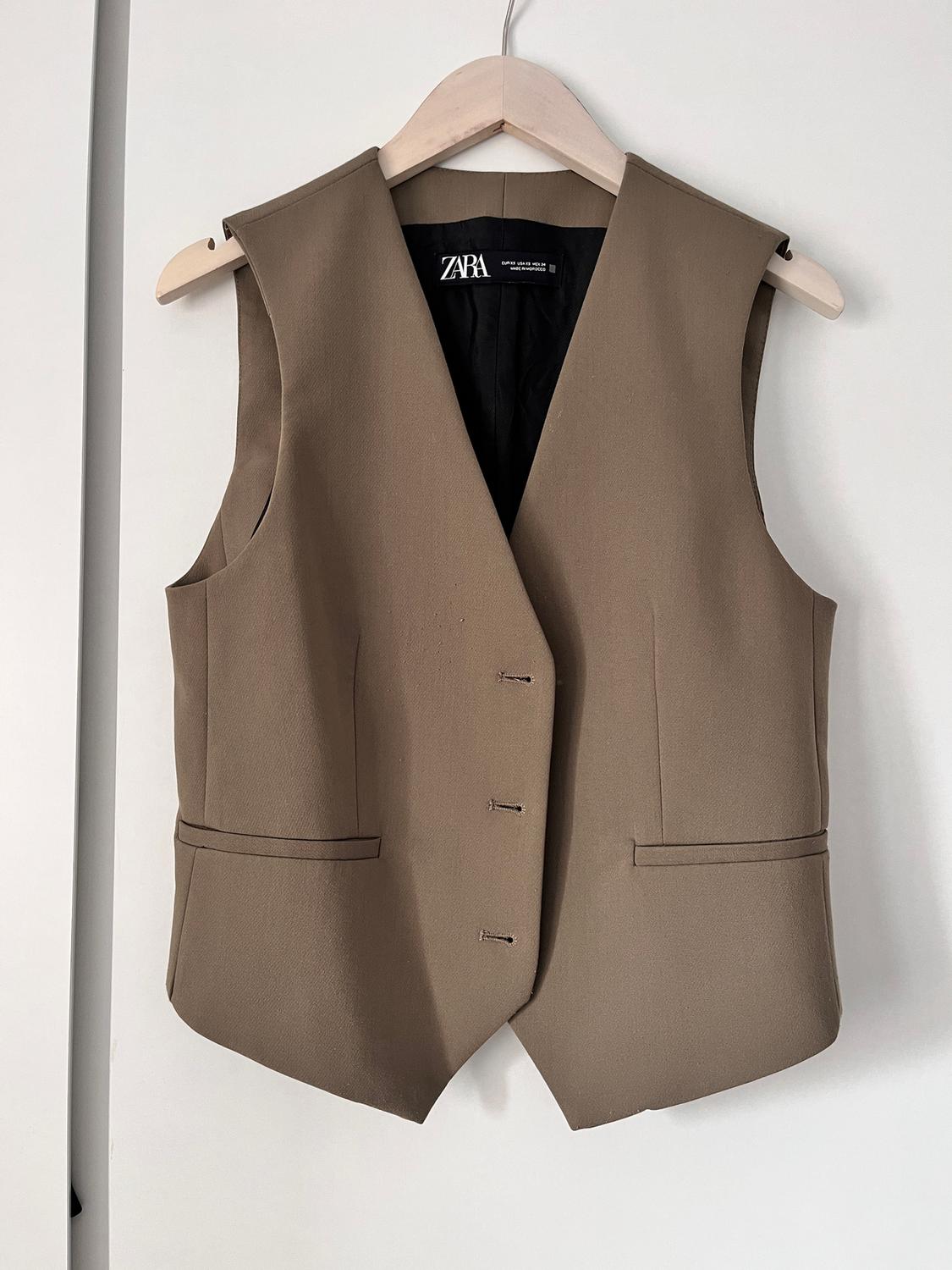 Brown sleeveless waistcoat hanging on a wooden hanger, with a visible Zara label inside. White background.
