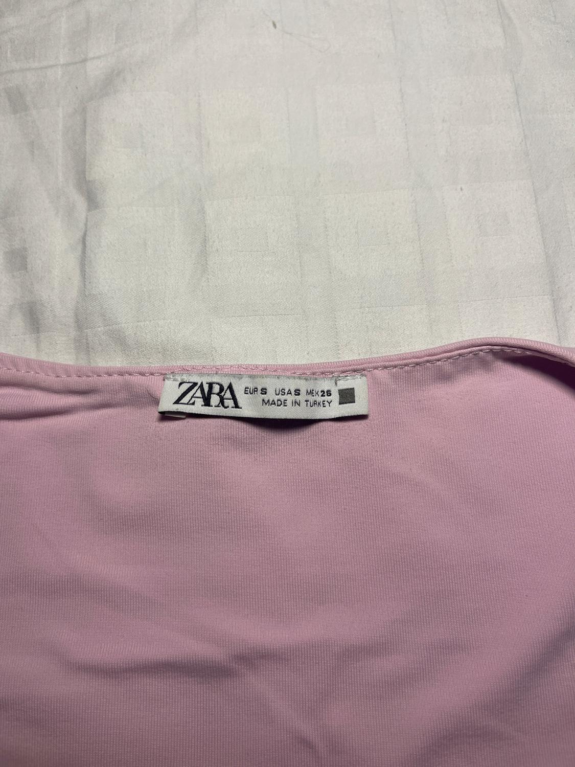 Ετικέτα ρούχων ZARA σε ροζ ύφασμα, αναγράφει μέγεθος EUR S USA S MX 26, κατασκευασμένο στην Τουρκία, σε λευκό φόντο.