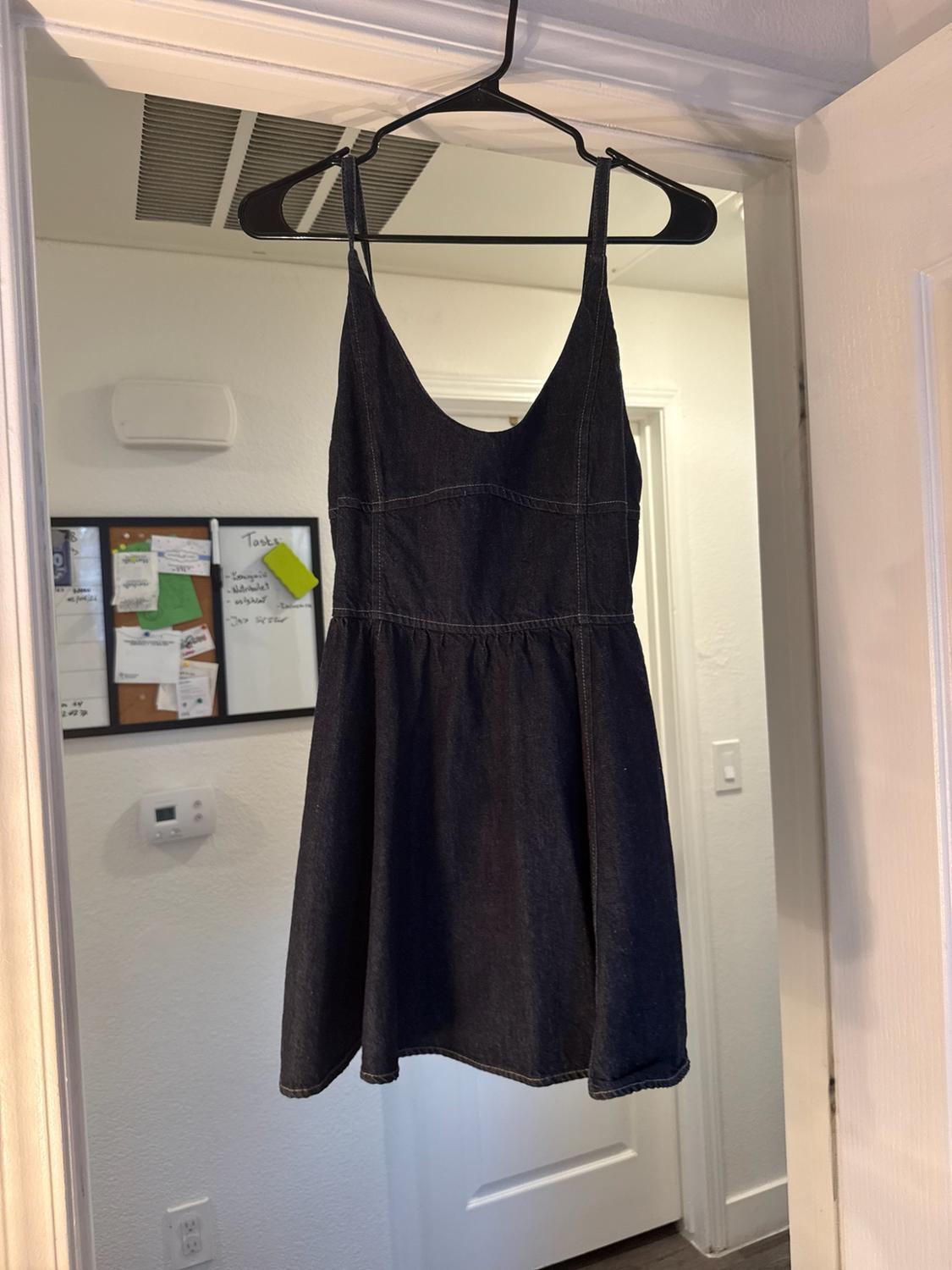 Dark denim spaghetti strap dress with visible stitching, hanging on a door. In the background, there is a white board with the word Tasks.