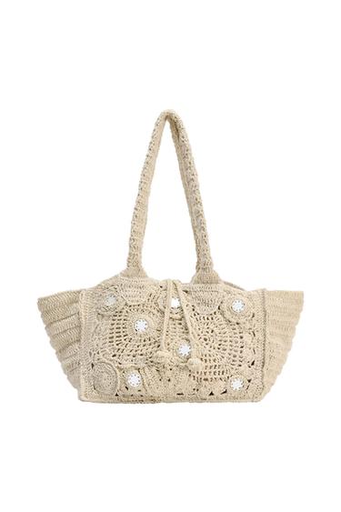 Beige crochet tote bag with handles, openwork circular motifs, drawstring with tassels, and mirror appliqués on a white background.