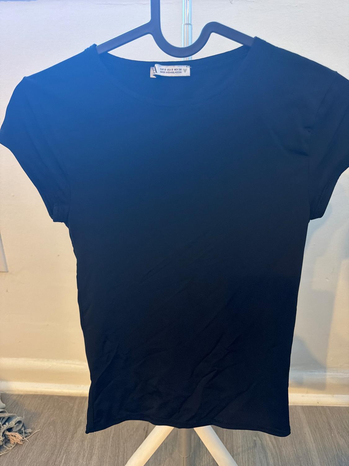 Black short-sleeved t-shirt on a black hanger in front of a white wall. Label: EUR S USA S MEX 26 MADE IN BANGLADESH.