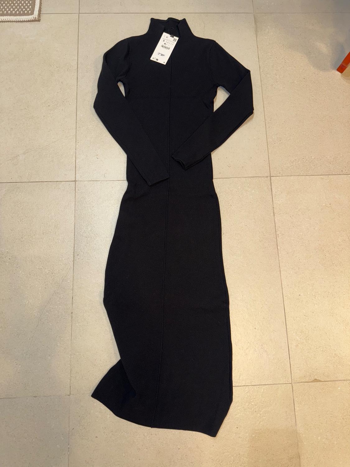 Black long-sleeved, high-neck dress laid out on a light tiled floor, with a visible price tag.