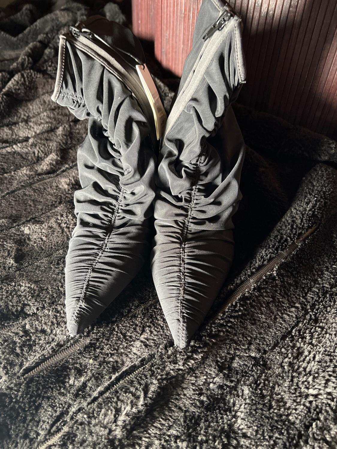 Black boots with ruching and zippers, on textured dark fabric. Soft light highlights design details.