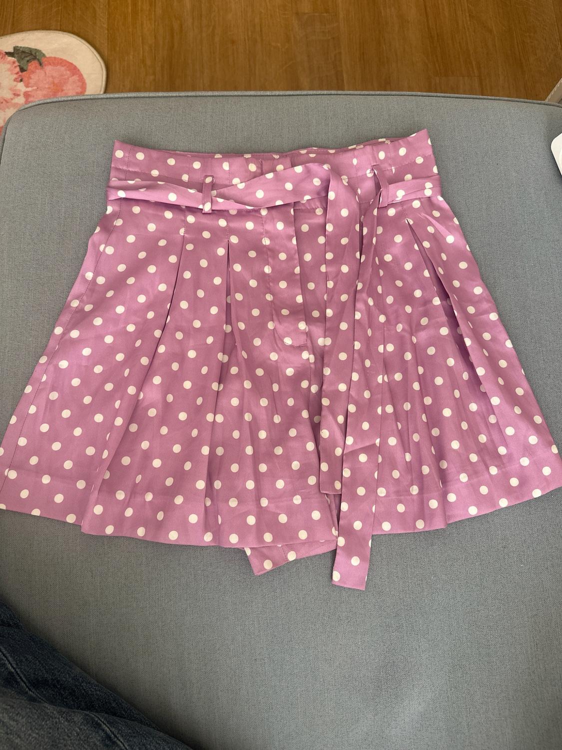 Lilac pleated shorts with white polka dots and a tie-knot belt. Relaxed composition on a grey seat with a wooden floor.