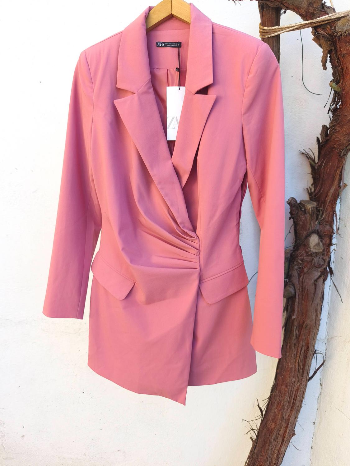 Pink blazer hanging on a wooden hanger, with visible tag. White background and tree branch on the right.