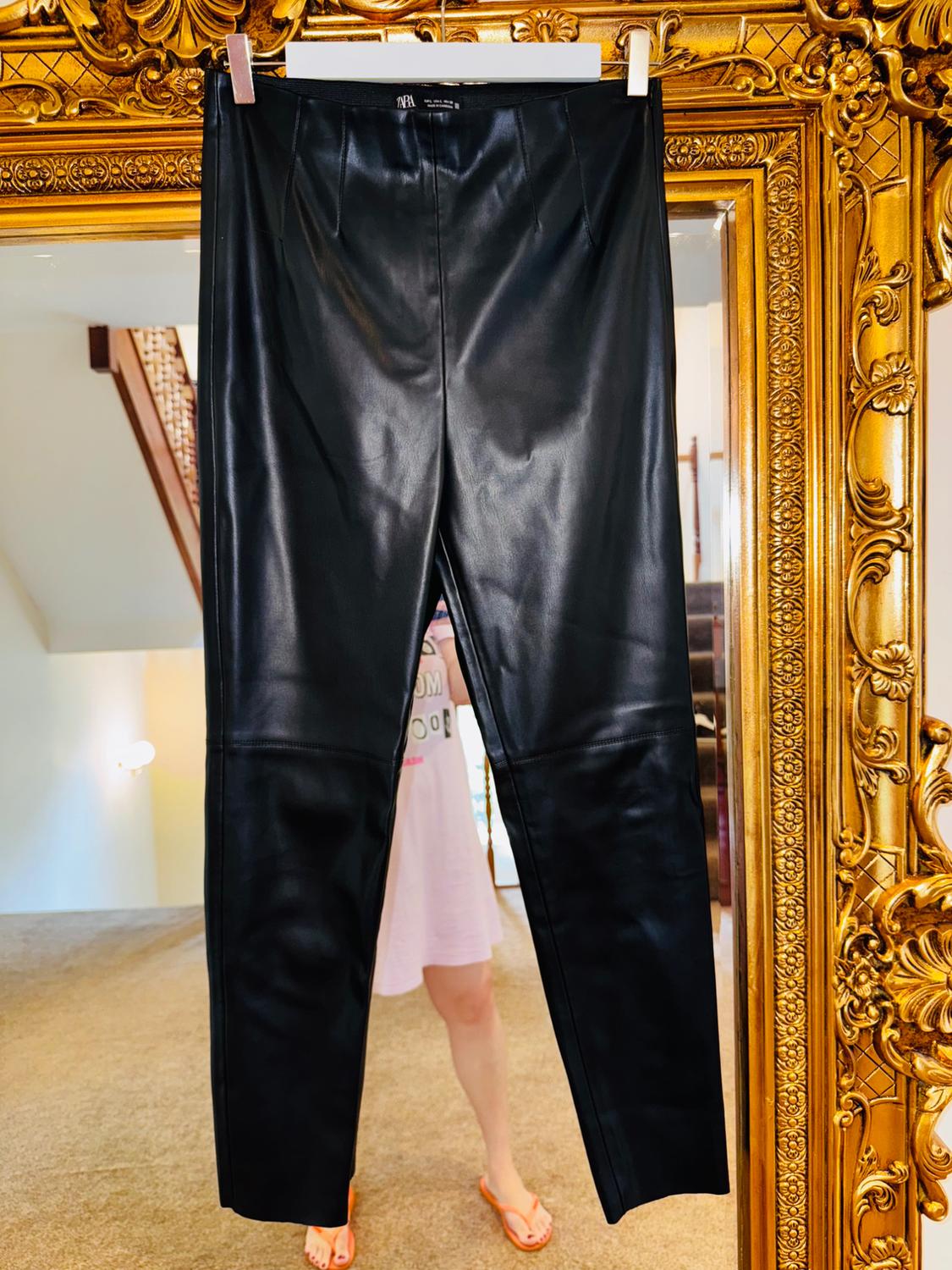 Black trousers hanging in an ornate gold frame. Reflection of a person in a pink dress and orange sandals in the mirror.