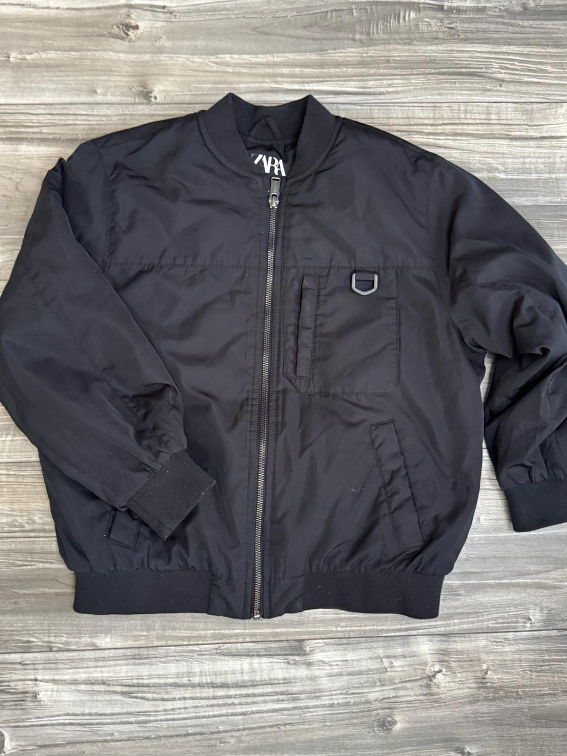 Black bomber jacket against a wooden background. Zip fastening, elasticated collar and cuffs. Pocket with ring on the chest. ZARA label.