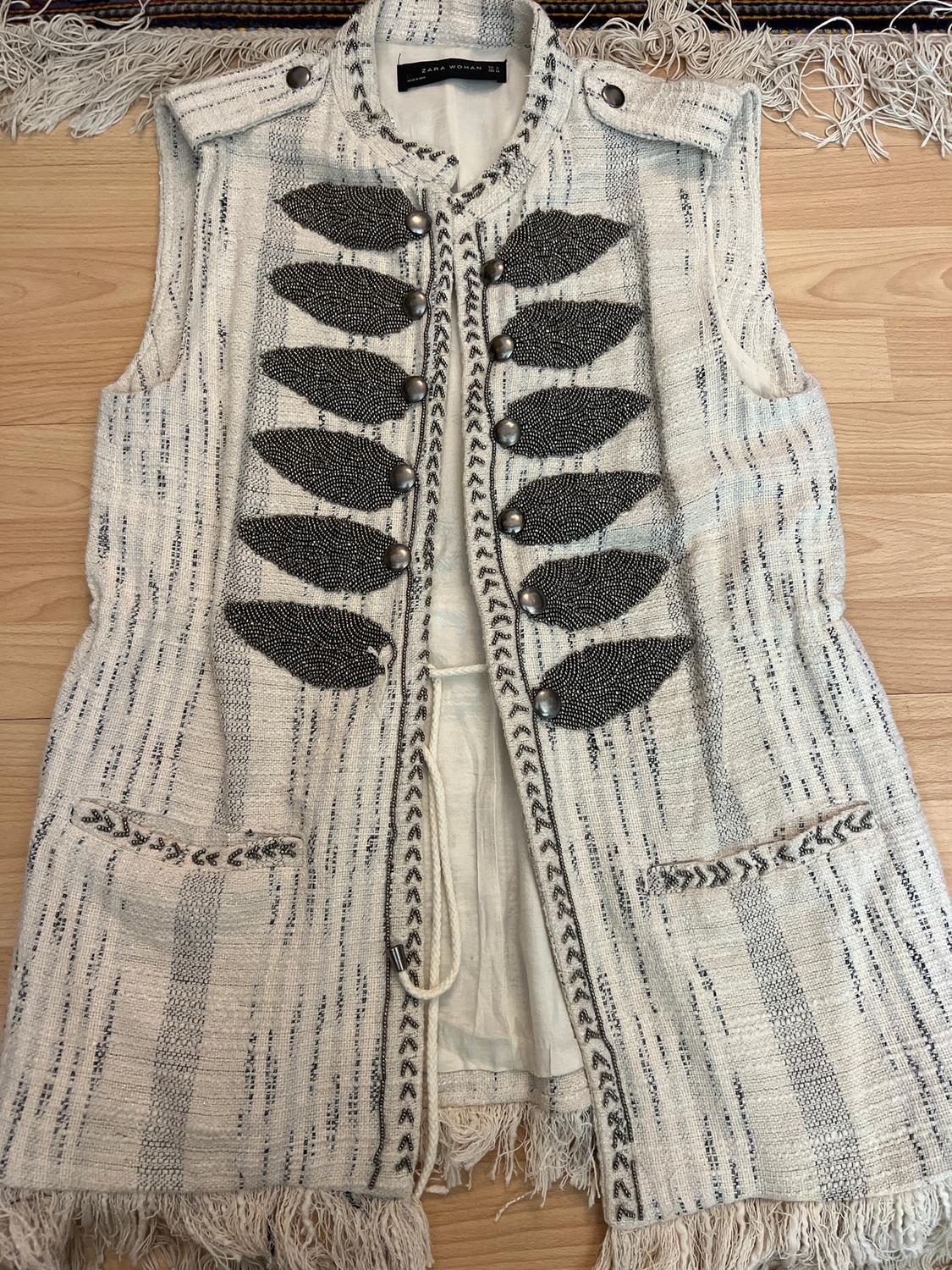 White textured sleeveless waistcoat with metallic button details and black embellishments, on a light wood background.