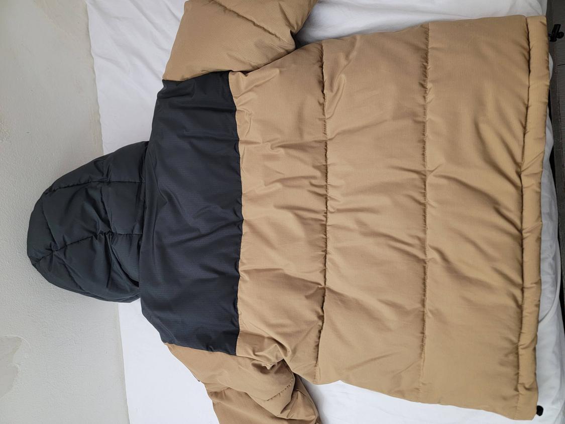 Quilted jacket with a hood, camel-colored with a black upper section, extended over a white fabric.