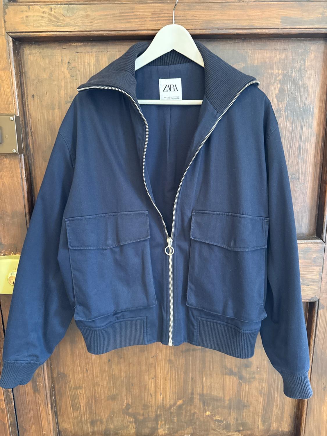 Blue jacket with a zip and large pockets, hanging on a white hanger in front of a wooden door. Visible label: ZARA.
