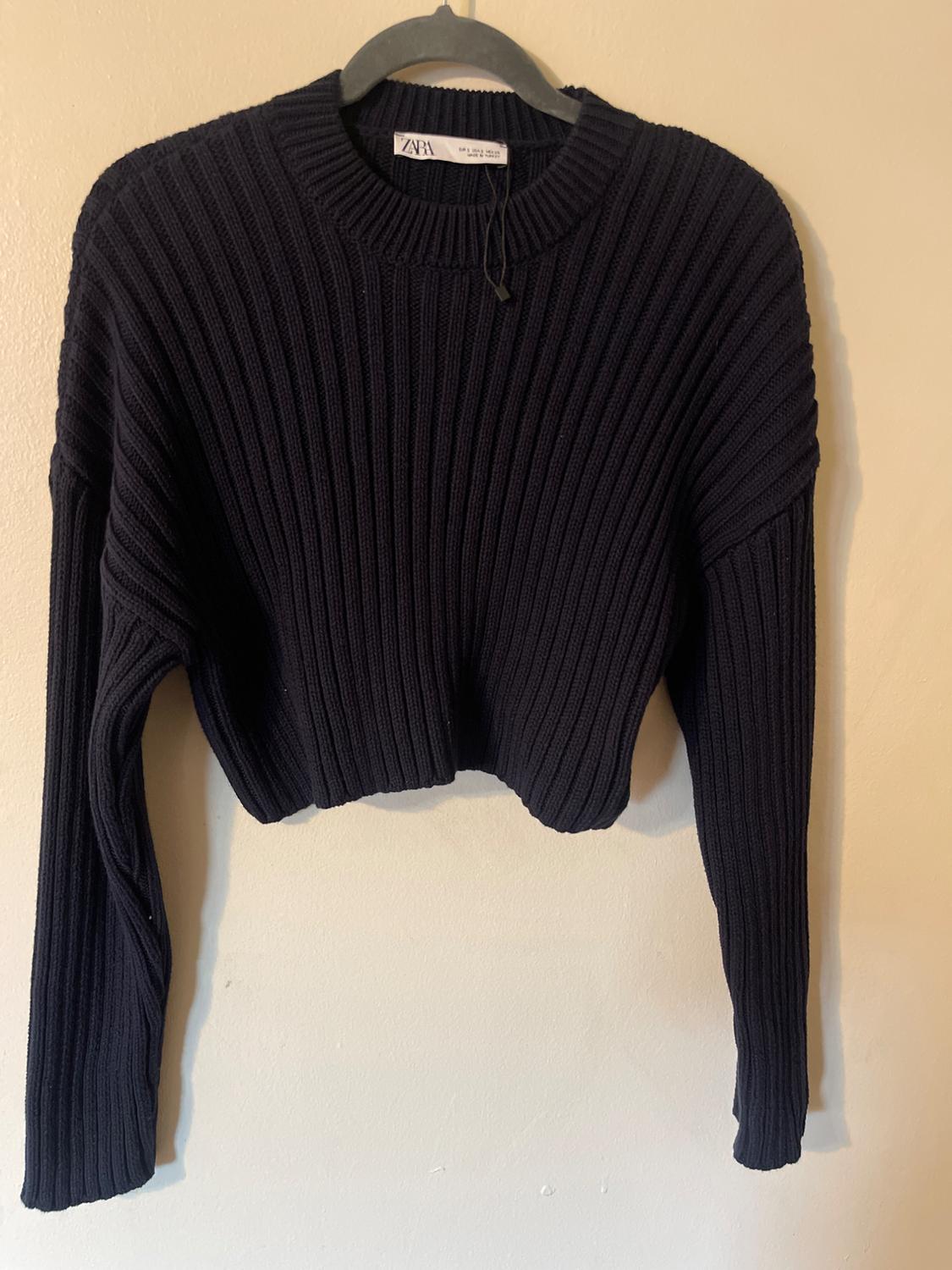 Black chunky knit sweater hanging on a black hanger. Visible tag with the text "ZARA". Beige wall background.