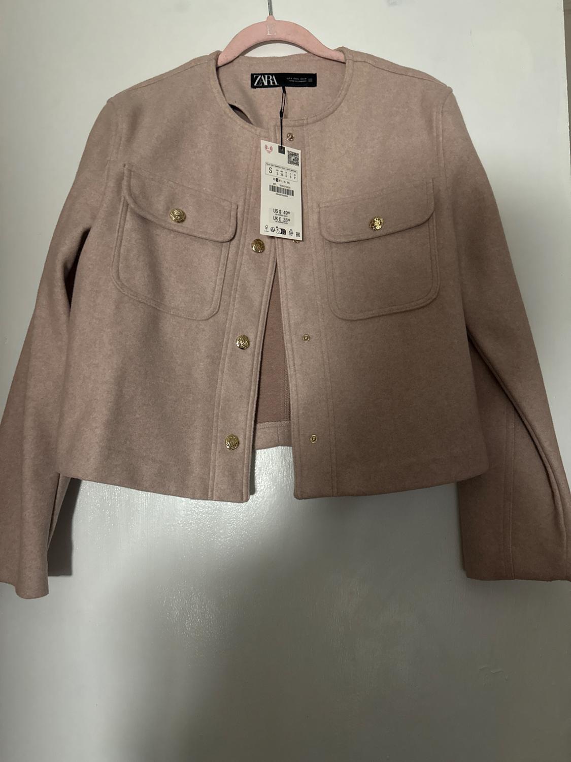 Short beige jacket with gold buttons and front pockets, hanging on a pink hanger. Visible tag with price and size.