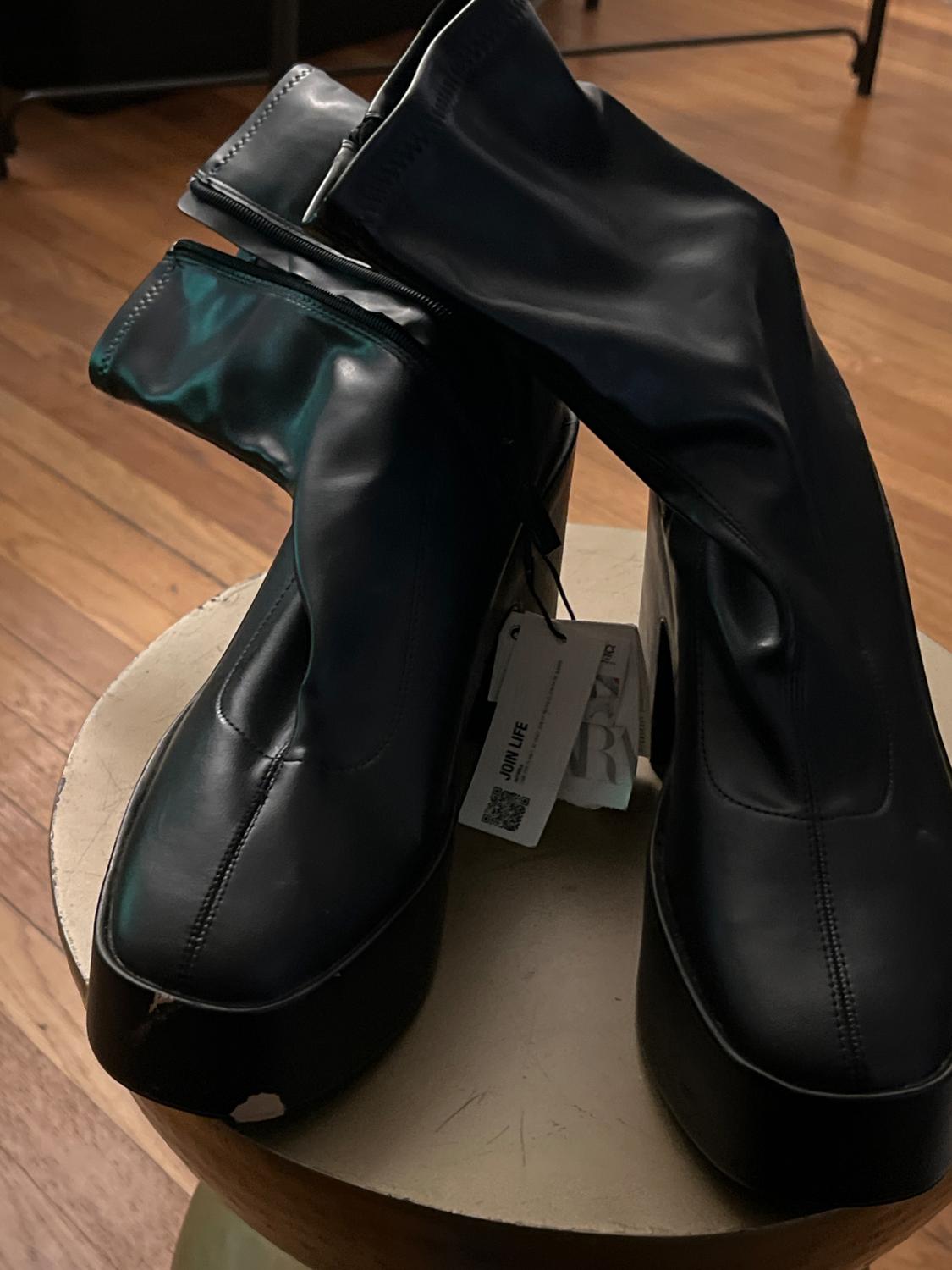 Black platform boots on a wooden table. Visible label with text "JOIN LIFE". Interior background of wooden flooring.