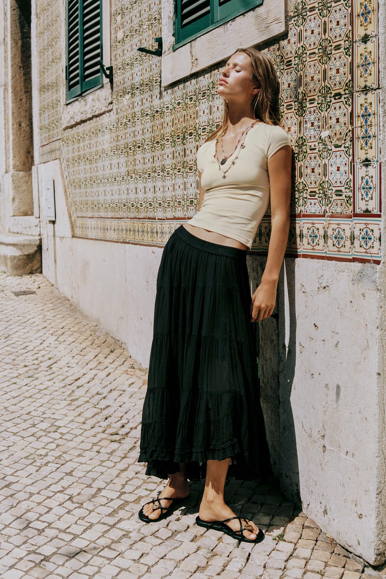 Beige short-sleeve t-shirt set, tiered black midi skirt with an elastic waistband and asymmetrical hem, and black strappy sandals.