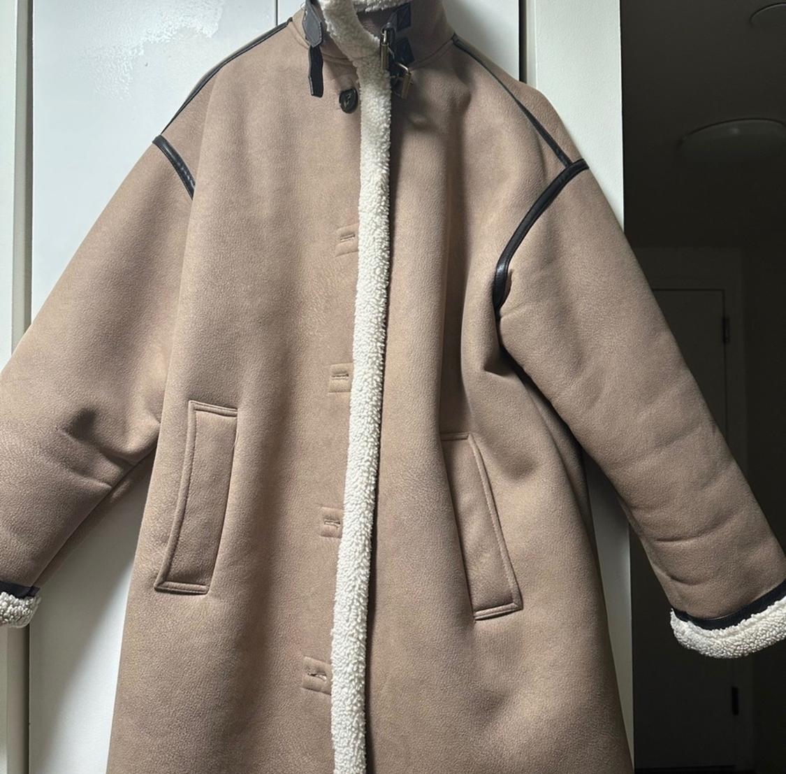 Beige coat hanging on a door, with black leather details on the shoulders and white wool trim on the closure and cuffs.