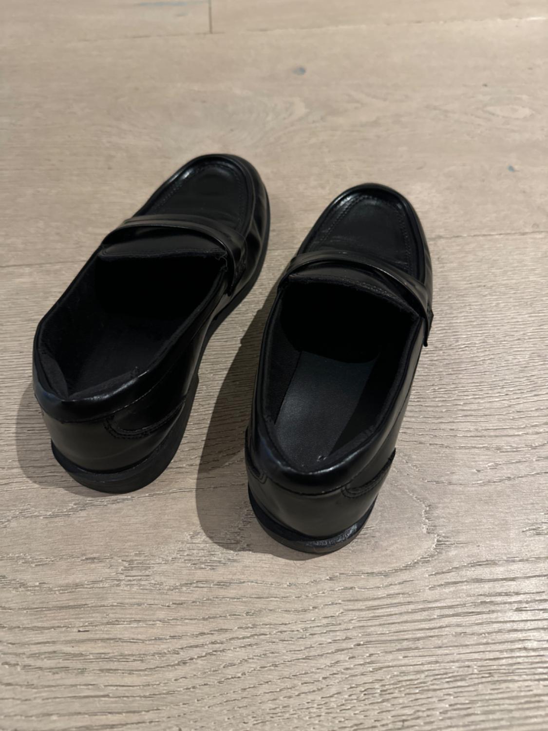 Black patent leather shoes on a light wooden floor, viewed from above, showcasing their classic and elegant design.