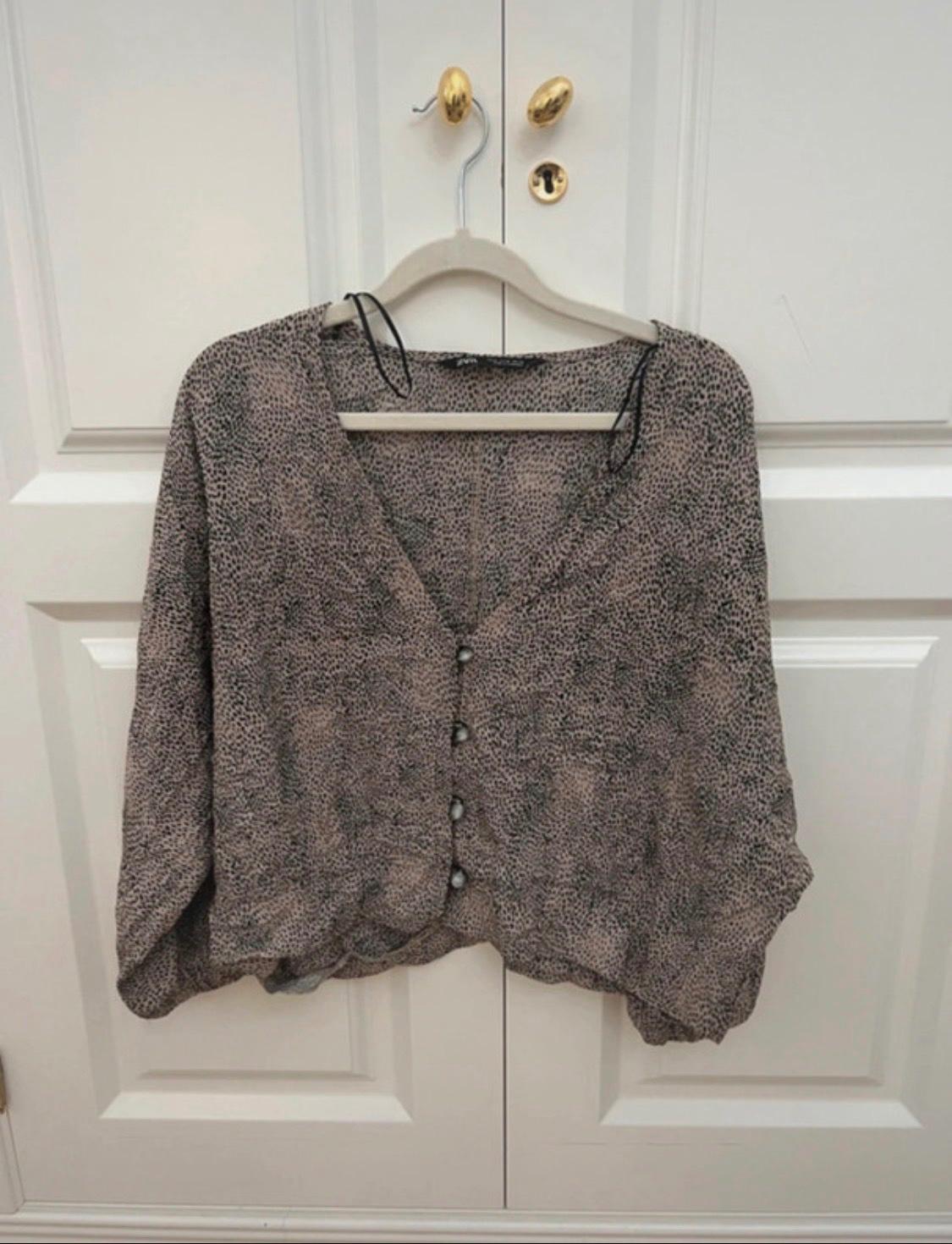 Beige blouse with black spot print, V-neck and front buttons, hanging on a hanger from the handle of a white door.